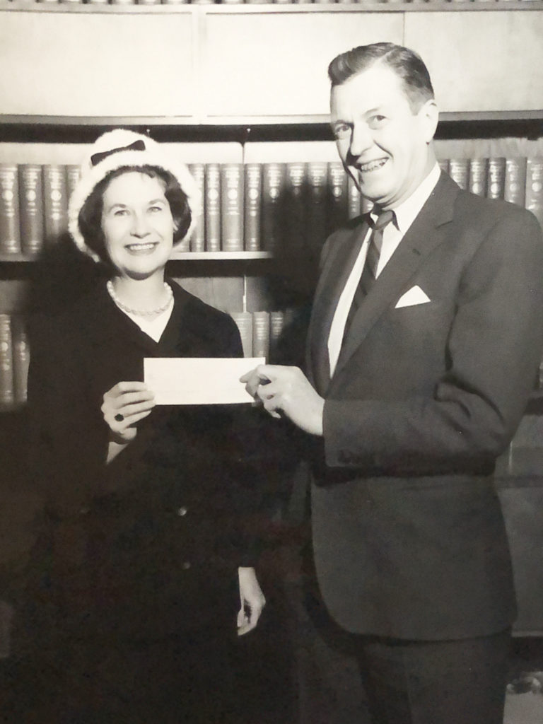 Two people standing in a library setting with a woman on the left and a man on the right. The woman is handing a check to the man and they are both smiling.