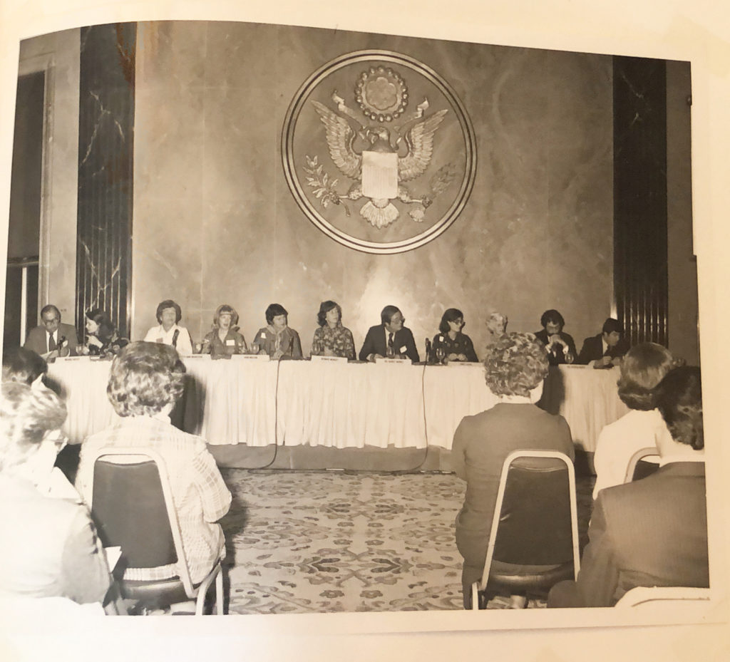 Multiple people seated at a long table as panel speakers. The view is facing the panel from the perspective of the audience.