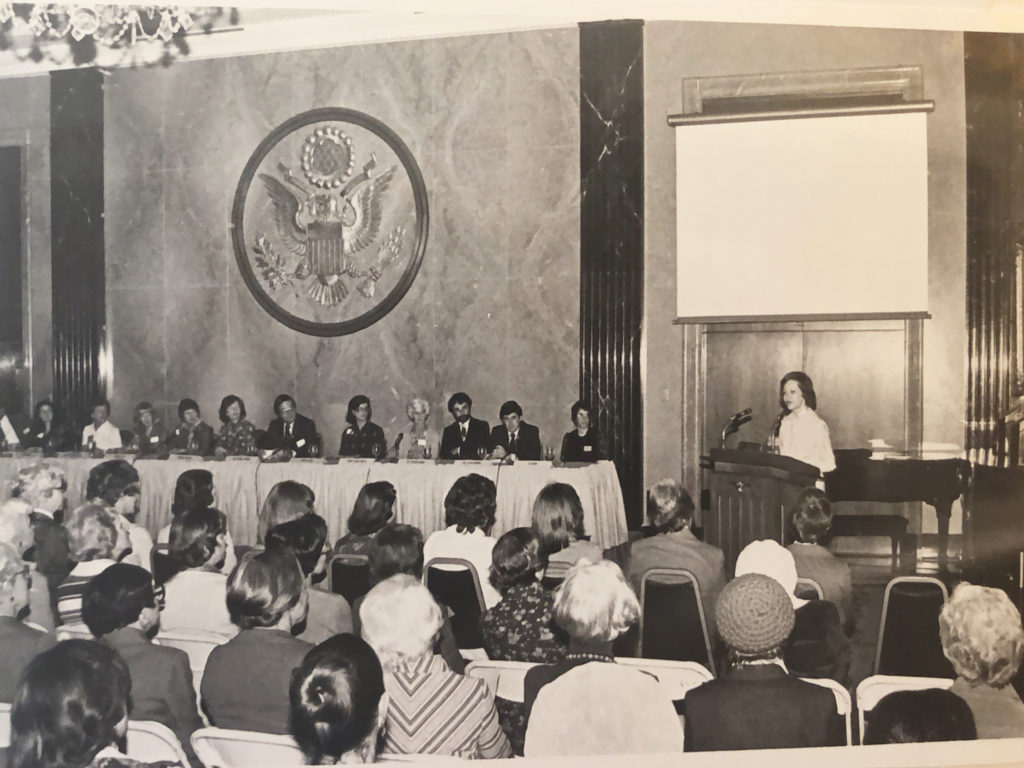 Rosalynn Carter is shown standing at a podium at the front of a large room. To her right is the long table with the panel members. The view is from the audience to Ms. Carter's left.